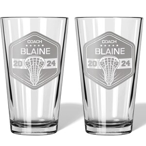 May include: Two clear glass pint glasses with a lacrosse-themed design. Each glass features a shield with the text "COACH BLAINE" above a lacrosse stick graphic and the numbers "20 24".