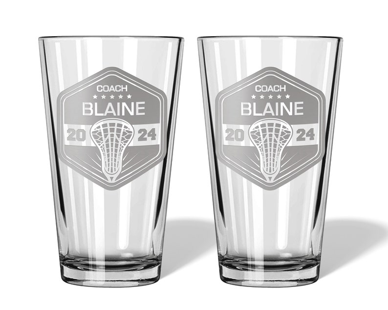 May include: Two clear glass pint glasses with a lacrosse-themed design. Each glass features a shield with the text "COACH BLAINE" above a lacrosse stick graphic and the numbers "20 24".