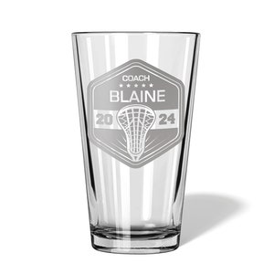 May include: A clear glass pint with a lacrosse-themed design. The design includes the word "COACH", the name "BLAINE", the year "2024", and a lacrosse stick and net graphic. The glass is suitable for beverages.
