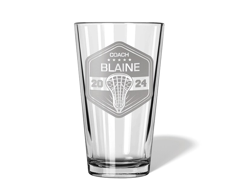 May include: A clear glass pint with a lacrosse-themed design. The design includes the word "COACH", the name "BLAINE", the year "2024", and a lacrosse stick and net graphic. The glass is suitable for beverages.
