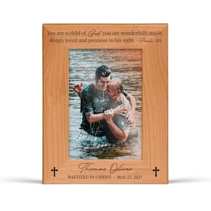 K&ouml;nnte beinhalten: Ein Holzfotorahmen mit einem Bild von zwei M&auml;nnern im Wasser, die sich umarmen. Der Rahmen hat Text oben und unten, darunter "You are a child of God" und "Thomas Oliver, Baptized in Christ, May 23, 2027". Kreuze sind auf dem Rahmen.