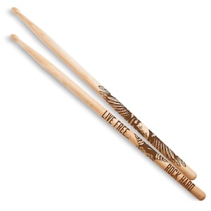 May include: Two wooden drumsticks with a natural finish. One stick features the text "LIVE FREE..." and the other reads "ROCK HARD." Both sticks have a brown eagle design. The drumsticks are angled and set against a white background.