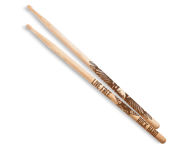 May include: Two wooden drumsticks with a natural finish. One stick features the text "LIVE FREE..." and the other reads "ROCK HARD." Both sticks have a brown eagle design. The drumsticks are angled and set against a white background.