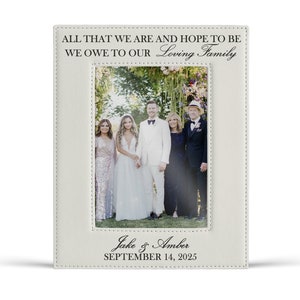 May include: A rectangular off-white leather picture frame with stitched edges. The top text reads ALL THAT WE ARE AND HOPE TO BE WE OWE TO OUR Loving Family. The bottom text reads Jake & Amber SEPTEMBER 14, 2025. A wedding photo is inside.