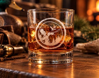 May include: A clear glass tumbler filled with amber liquid and ice cubes. The front of the glass features a white etched circular design depicting a flying duck above a forest silhouette, framed by a textured ring resembling tree bark.