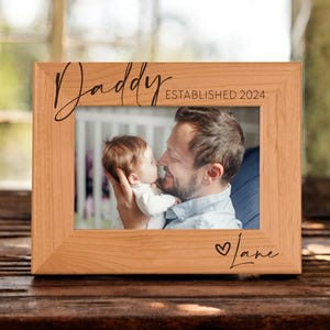 May include: A wooden photo frame with the text "Daddy Established 2024" and a heart with the name "Lane" engraved on the bottom. The frame has a photo of a father and child.