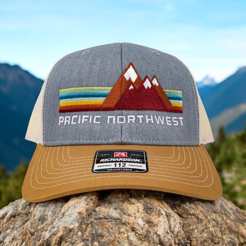May include: A gray and beige trucker hat with a red, orange, yellow, and blue mountain range design and the text "PACIFIC NORTHWEST". The hat has a brown adjustable strap with the text "RICHARDSON 112 ORIGINAL TRUCKER ADJUSTABLE".