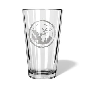 May include: A clear glass pint glass with a design etched into the side. The design is a circle with a tree trunk texture, a bird flying over a forest, and a deer in the forest.