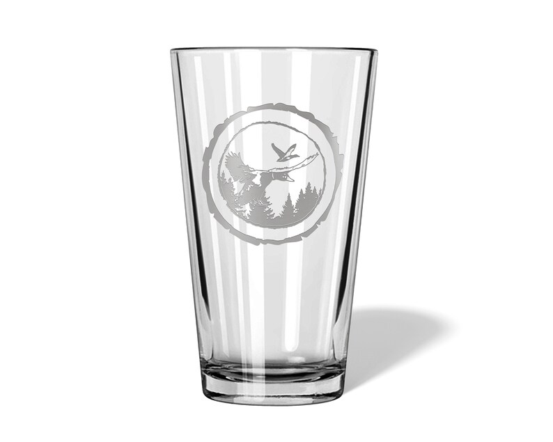 May include: A clear glass pint glass with a design etched into the side. The design is a circle with a tree trunk texture, a bird flying over a forest, and a deer in the forest.