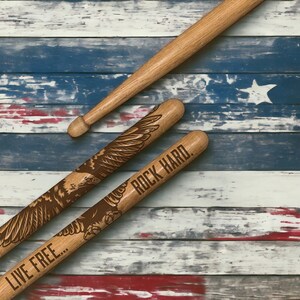 May include: Three wooden drumsticks, two with engraved eagle designs and the phrases "LIVE FREE..." and "ROCK HARD." The drumsticks are set against a backdrop of a distressed American flag.