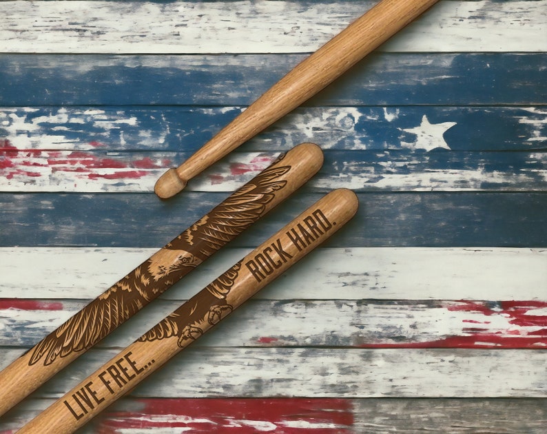 May include: Three wooden drumsticks, two with engraved eagle designs and the phrases "LIVE FREE..." and "ROCK HARD." The drumsticks are set against a backdrop of a distressed American flag.