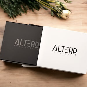 May include: A black and white box with the logo "ALTERD INDUSTRIES" printed on both sides. The box is on a wooden surface.