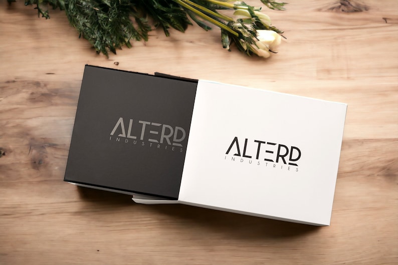 May include: A black and white box with the logo "ALTERD INDUSTRIES" printed on both sides. The box is on a wooden surface.