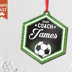 May include: A hexagonal wooden ornament with a green grass background. The ornament features a black and white soccer ball with a white border and a black star on each side. The text "2020 COACH James" is printed in white on a black background.