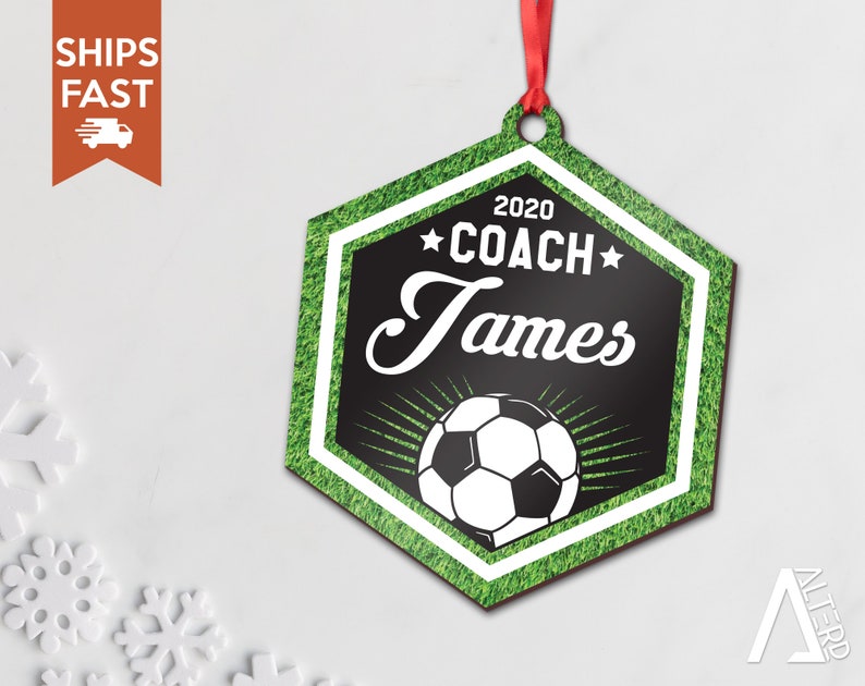 May include: A hexagonal wooden ornament with a green grass background. The ornament features a black and white soccer ball with a white border and a black star on each side. The text "2020 COACH James" is printed in white on a black background.