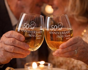 May include: Two clear stemless wine glasses filled with a golden liquid, each etched with the text 50th ANNIVERSARY, the names Ned & Karen, and the date 9.9.89, featuring a decorative swirl design above the anniversary text.