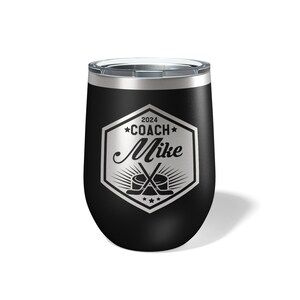 May include: A black stainless steel tumbler with a clear lid, featuring a silver laser-engraved hexagonal emblem. The design includes the text 2024, COACH, and Mike, accompanied by a graphic of two crossed hockey sticks and a hockey puck.
