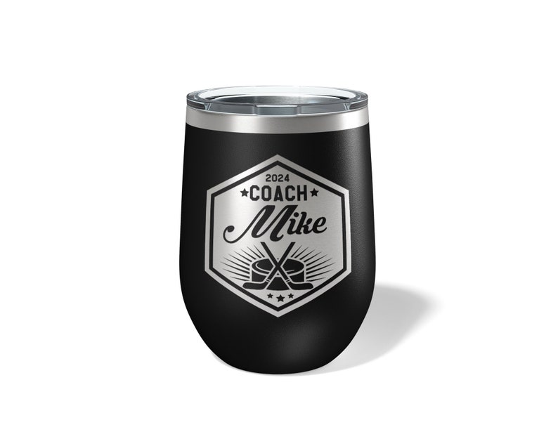 May include: A black stainless steel tumbler with a clear lid, featuring a silver laser-engraved hexagonal emblem. The design includes the text 2024, COACH, and Mike, accompanied by a graphic of two crossed hockey sticks and a hockey puck.