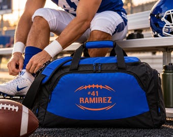 May include: A black and blue duffel bag featuring an orange graphic of an American football with the text #41 RAMIREZ printed in the center. The bag has black handles, a shoulder strap, and multiple zippered compartments for storage.