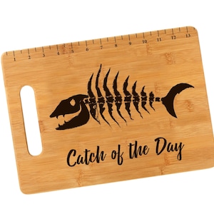 K&ouml;nnte beinhalten: Ein braunes Bambus-Schneidebrett mit einem Fischskelett-Design und dem Text "Catch of the Day". Das Brett hat einen Griff und ein Lineal, das auf der Oberkante aufgedruckt ist.