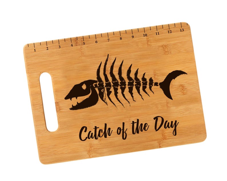 K&ouml;nnte beinhalten: Ein braunes Bambus-Schneidebrett mit einem Fischskelett-Design und dem Text "Catch of the Day". Das Brett hat einen Griff und ein Lineal, das auf der Oberkante aufgedruckt ist.