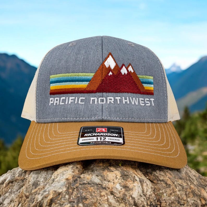May include: A gray and beige trucker hat with a red, orange, yellow, and blue mountain range design and the text "PACIFIC NORTHWEST". The hat has a brown adjustable strap with the text "RICHARDSON 112 ORIGINAL TRUCKER ADJUSTABLE".