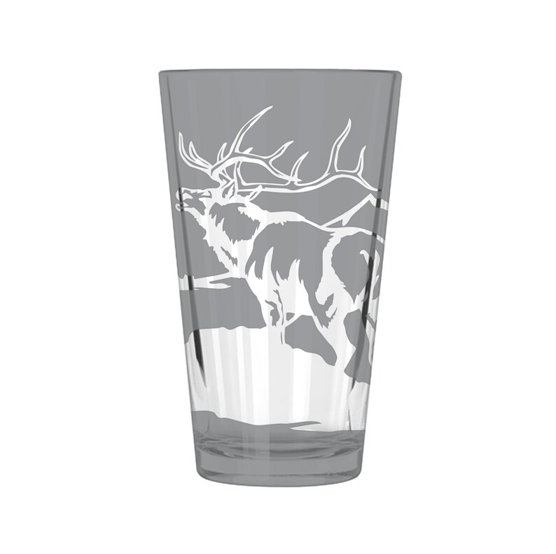 Puede incluir: Un vaso de vidrio transparente con un tinte gris presenta un dise&ntilde;o grabado en blanco de un alce de pie en un paisaje de monta&ntilde;a.