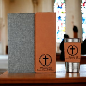 May include: A gray and brown leatherette journal with a cross and wreath design and the text "2 Timothy 4:2 Pastor Tracy W. Wilson". A matching brown leatherette tumbler with the same design and text is also shown.
