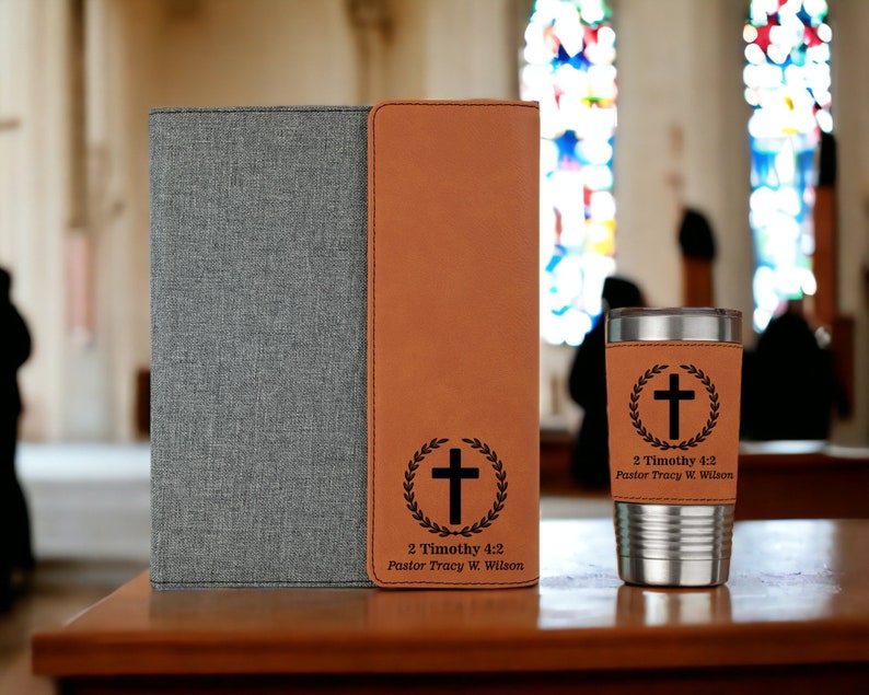 May include: A gray and brown leatherette journal with a cross and wreath design and the text "2 Timothy 4:2 Pastor Tracy W. Wilson". A matching brown leatherette tumbler with the same design and text is also shown.
