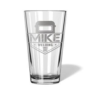 May include: A clear pint glass with a custom design. The design includes a welding mask graphic above the name "MIKE" and the words "WELDING SINCE 1997" in a diamond-shaped emblem. The glass is suitable for beverages.