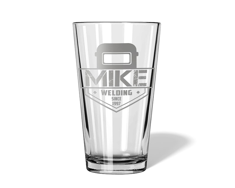 May include: A clear pint glass with a custom design. The design includes a welding mask graphic above the name "MIKE" and the words "WELDING SINCE 1997" in a diamond-shaped emblem. The glass is suitable for beverages.