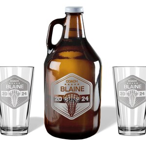 May include: A brown glass growler and two clear pint glasses. Each glass and the growler feature a silver emblem with the text "COACH BLAINE 2024" and a lacrosse stick design. The growler has a white cap.