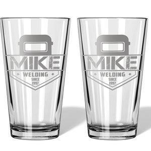May include: Two clear pint glasses with a personalized design. Each glass features a welding helmet graphic above the name "MIKE" and the words "WELDING SINCE 1997" in a diamond-shaped emblem. The glasses are ideal for a welder.