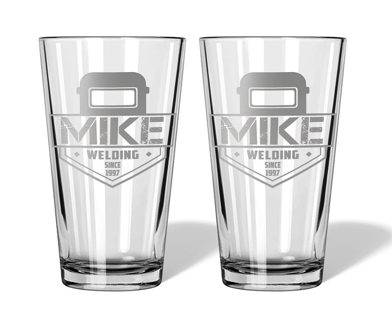 May include: Two clear pint glasses with a personalized design. Each glass features a welding helmet graphic above the name "MIKE" and the words "WELDING SINCE 1997" in a diamond-shaped emblem. The glasses are ideal for a welder.
