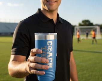May include: A blue stainless steel tumbler with a silver rim and a laser-engraved design featuring a soccer ball icon and the text "COACH" in a bold, block-style font. The tumbler has a textured exterior with horizontal ridges near the base.