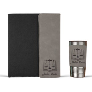 May include: A black notebook with a gray vertical accent strip and a matching gray tumbler. Both items feature a black engraved graphic of a scale of justice over an open book, with the name Jackson Lourie printed in a cursive script font below.