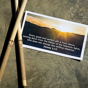 May include: Two light-coloured wooden drumsticks with ribbed grips and a cross symbol above the text 1:17, positioned next to a rectangular card featuring a sunset landscape and the text: Every good and perfect gift is from above, coming down from the Father of the heavenly lights, who does not change like shifting shadows. James 1:17.