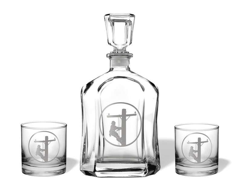 May include: A clear glass decanter with a matching stopper and two matching lowball glasses. Each piece features an etched circular design depicting a utility worker climbing a wooden power pole, rendered in a frosted, matte finish.