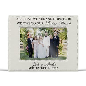 May include: A rectangular off-white leatherette picture frame featuring stitched borders. The top text reads ALL THAT WE ARE AND HOPE TO BE WE OWE TO OUR Loving Parents. The bottom text reads Jake & Amber SEPTEMBER 14, 2025.