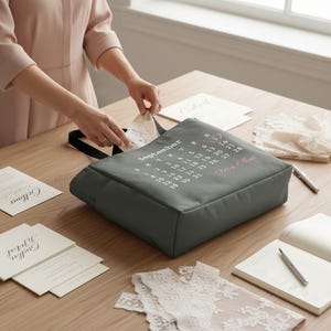 May include: A grey tote bag with a September calendar design and the text "Mrs. Elliott." The bag has a black handle and is surrounded by wedding stationery and lace, suggesting a bridal theme.