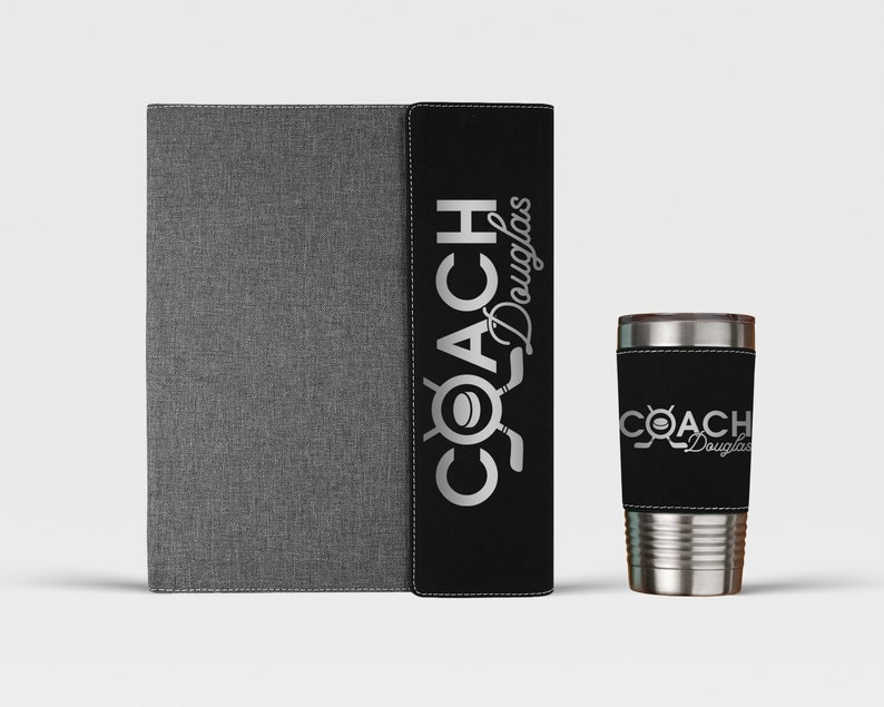 May include: A black and gray notebook with a black and silver "COACH Douglas" logo on the cover. Next to the notebook is a silver tumbler with a black and silver "COACH Douglas" logo on the sleeve.