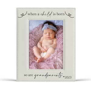 Pu&ograve; includere: Cornice fotografica color crema con il testo "when a child is born" e "so are grandparents 2023." La cornice contiene una foto di un neonato avvolto in una coperta rosa.