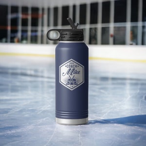 May include: A navy blue insulated water bottle with a black lid and straw. The front features a white hexagonal graphic with the text 2024 COACH Mike, accompanied by an illustration of two crossed hockey sticks and a puck centered below the name.