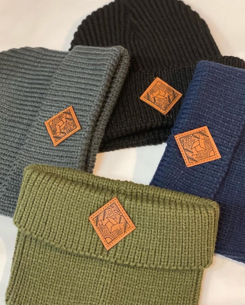 Puede incluir: Cuatro gorros de punto en diferentes colores, negro, gris, verde y azul, con un parche de cuero que presenta un dise&ntilde;o de ciervo estilizado en cada uno.