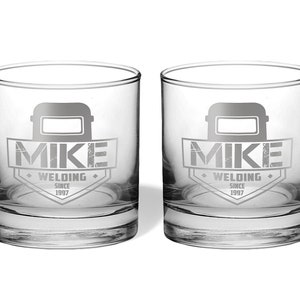 May include: Two clear, round-bottomed drinking glasses. Each glass features a gray graphic of a welding mask above the name "MIKE" and the words "WELDING SINCE 1997" in a shield design. The glasses are on a white background.