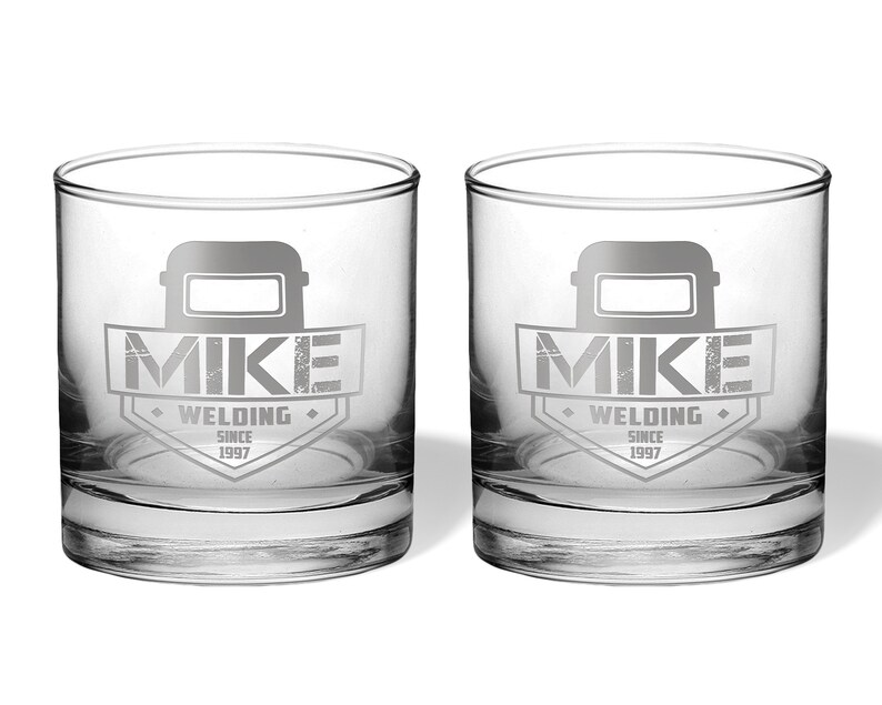 May include: Two clear, round-bottomed drinking glasses. Each glass features a gray graphic of a welding mask above the name "MIKE" and the words "WELDING SINCE 1997" in a shield design. The glasses are on a white background.