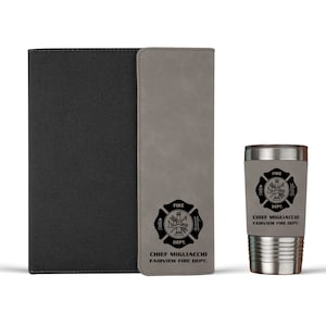 Pu&ograve; includere: Un taccuino nero con un pannello verticale grigio e un bicchiere in acciaio inossidabile grigio abbinato. Entrambi gli articoli presentano un emblema nero dei vigili del fuoco con il testo "CHIEF MIGLIACCIO FAIRVIEW FIRE DEPT." sulle superfici grigie.