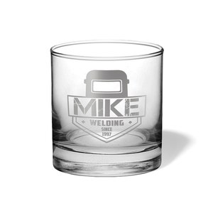 May include: A clear, engraved rocks glass featuring the name "MIKE" above the word "WELDING" and the year "SINCE 1997". The design includes a welding mask graphic. The glass is cylindrical and sits on a white surface.