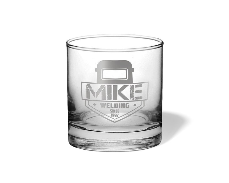 May include: A clear, engraved rocks glass featuring the name "MIKE" above the word "WELDING" and the year "SINCE 1997". The design includes a welding mask graphic. The glass is cylindrical and sits on a white surface.