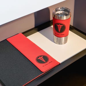 K&ouml;nnte beinhalten: Ein schwarzer und roter Lederordner mit einem medizinischen Symbol und dem Text "Dr. Dara Labron" auf der Vorderseite. Eine Edelstahl-Reisebecher mit einer roten Lederh&uuml;lle und dem gleichen medizinischen Symbol und Text befindet sich auf einer wei&szlig;en Oberfl&auml;che.